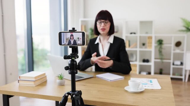 Caucasian adult woman wearing professional attire recording video blog about business topics using smartphone camera in modern office environment. Includes elements like notebook, laptop. - Powered by Adobe
