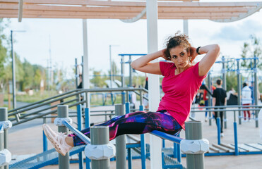 With focused determination, a woman does sit-ups on an outdoor exercise bar, surrounded by a lively park atmosphere.