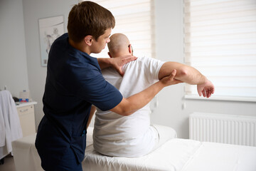 Fototapeta premium Chiropractor adjusting male patient’s shoulder in clinic. Physiotherapist performing manual therapy to relieve pain, improve mobility, support rehabilitation after injury or musculoskeletal disorder.