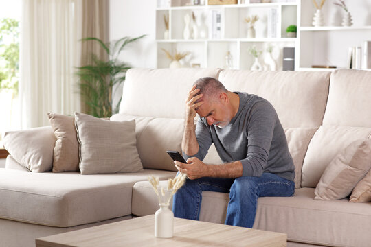 Worried senior man checking bad news on phone at home - Powered by Adobe
