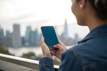 Woman holding smartphone against urban skyline at sunset. Blank phone screen ideal for mockup, app presentation, or social media design concept.