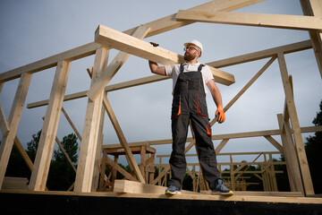 Construction worker carrying wooden beam on shoulder at building site. Carpenter in uniform and...