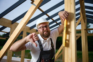 Frustrated construction worker showing thumbs down at building site. Disappointed builder in hard hat holding level tool, expressing failure, mistake, or poor construction result.