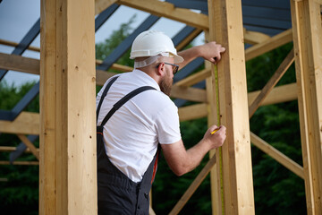 Construction worker measuring wooden beam with tape measure at building frame. Builder in safety helmet working on wooden house construction site outdoors.