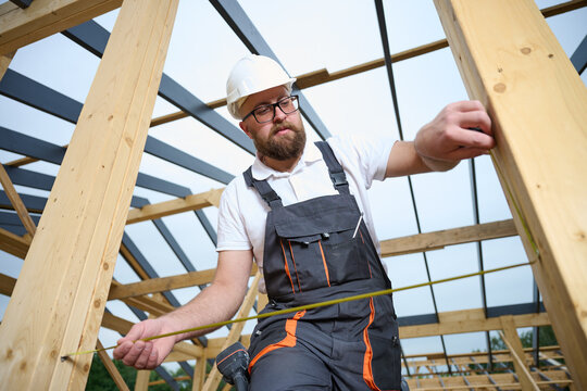 Construction worker measuring wooden beam with tape measure at building frame. Builder in safety helmet working on wooden house construction site outdoors.