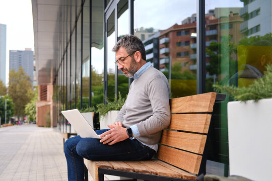 side view of Mature man working remote with laptop on city bench in barcelona