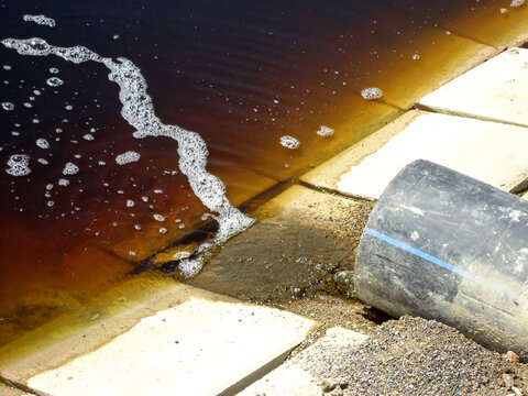 Detailed photo of heavily polluted water landfill leachate draining from pipe into evaporation pond with tiled edges, showing waste management, environmental contamination, industrial water treatment