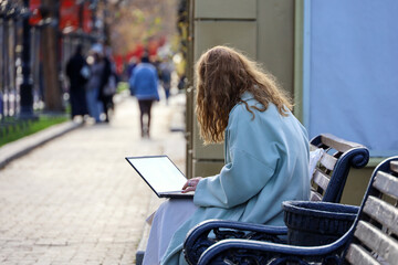 Young woman sitting with laptop on wooden bench on city street. Working in autumn park