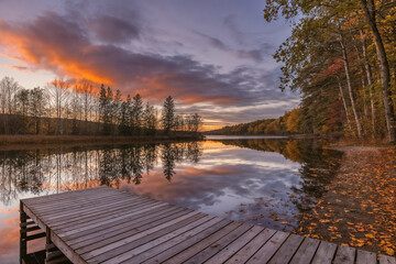 A low-angle, wide shot of a wooden dock extending into a calm lake, perfectly reflecting the dramatic autumn sunset sky with fiery orange and purple clouds, surrounded by trees with fall foliage.