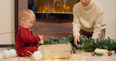 A young boy enjoys playing with colorful Christmas baubles and twinkling tree lights as his mother...