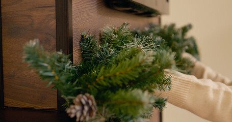 Hands carefully position a decorative fir branch with natural pinecones over a cozy fireplace, creating a festive holiday atmosphere in a warm setting.