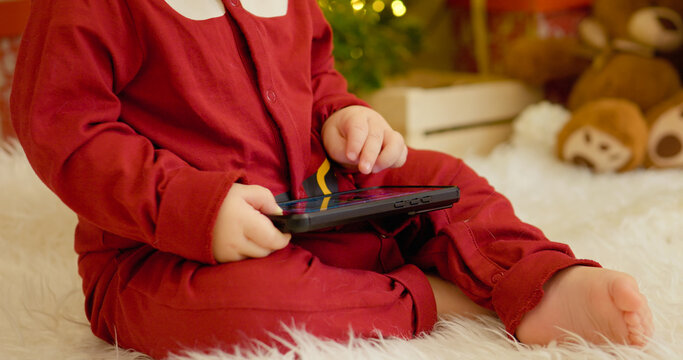A young boy sits on the floor, fully engaged with his smartphone amid a beautifully decorated Christmas tree and colorful ornaments, creating a festive atmosphere. - Powered by Adobe