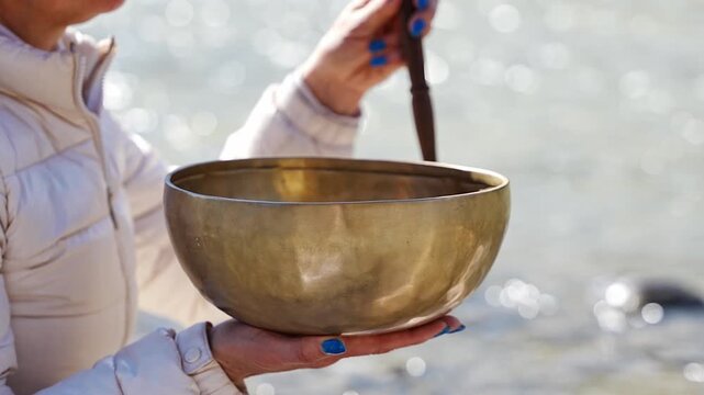 Close-up of hands playing a Tibetan singing bowl by a mountain river. Concept of sacred vibration, healing through touch, and meditative connection to natural energy