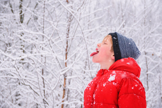Profile portrait of a young boy in a puffer jacket and woolly hat standing the woods with his tongue sticking ou catching snowflakes 