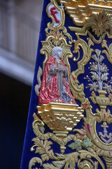Embroidered banner with a detailed figure of a young saint in red and blue robes, surrounded by ornate gold motifs on deep blue velvet. Granada, devotion