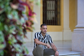 A young, handsome man with a beard, wearing a striped shirt and cargo trousers, is sitting on a...