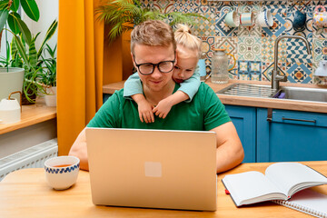 Father works remotely in the kitchen on a laptop, daughter little child distracts attention...