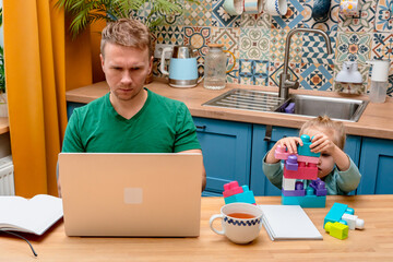 Father works remotely in the kitchen on a laptop, daughter little child plays with a construction...