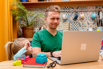 Father works remotely in the kitchen on a laptop, daughter little child distracts attention...