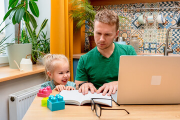 Father works remotely in the kitchen on a laptop, daughter little child distracts attention...