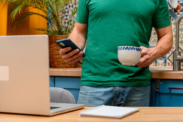 A young man works from home in the kitchen using a laptop. Freelancer working from home