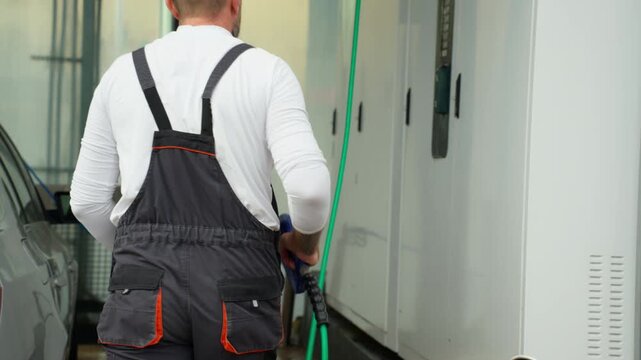 Man in overalls inserting credit card into payment terminal at self-service car wash before cleaning vehicle with high-pressure water gun