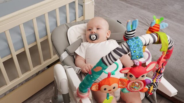 Peacefully playing baby lies in comfortable rocking chair with toys for entertainment, sucking on pacifier. Infant enjoys quiet play surrounded by soothing rattles, feeling safe - Powered by Adobe