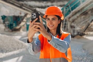 Smiling beautiful woman wearing safety hard hat and orange vest taking photo, holding mobile phone