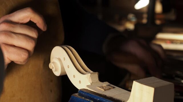 Extreme close-up of a luthier&rsquo;s hands refining the scroll and peg box with a hand tool, highlighting careful wood shaping, fine detail control, and secure vice positioning.