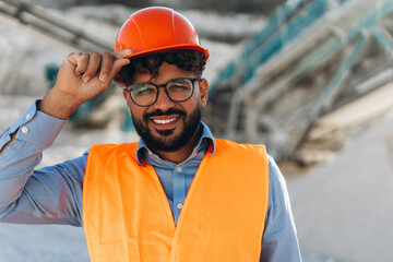 Smiling confident Middle Eastern engineer wearing glasses adjusting safety helmet at quarry site