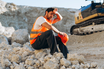 Tired male worker wearing orange safety vest sitting on rock, wiping sweat, resting near excavator © Maria Vitkovska