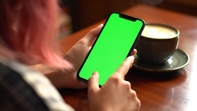 Closeup of a womans hands holding a smartphone with a green screen tapping and swiping while sitting at a table with a coffee cup in a cafe or home setting ideal for app promotion and digital content. - Powered by Adobe