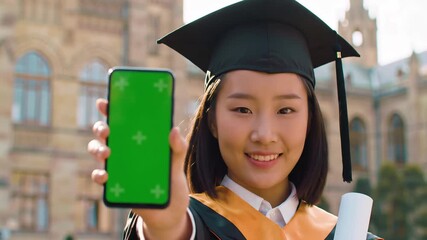 Happy Asian female graduate in academic dress and cap holding a smartphone with a green screen and a diploma smiling at the camera in front of a university building celebrating her academic achieveme.
