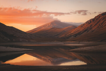 A breathtaking landscape shot captures a deep valley lake reflecting the surrounding mountains and the vibrant orange, pink, and red hues of a dramatic sunset or sunrise sky.