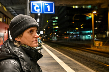 Woman in a black winter jacket and beanie waits on a night platform in Zurich