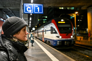 Woman in a black winter jacket and beanie waits on a night platform in Zurich. A train stands beside her under bright station lights in an urban setting