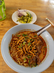 Authentic Northern Thai Khao Soi with tender chicken drumstick, rich coconut curry broth, and crispy noodles, served with traditional pickled mustard greens and fresh lime on a rustic wooden table in