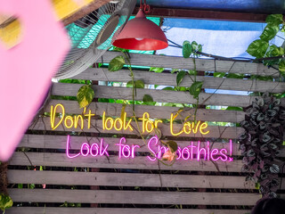 Vibrant neon sign illuminates a quirky message, 'Don't look for Love Look for Smoothies!', against a rustic wooden wall adorned with lush foliage and a soft-glowing red lamp, creating a relaxed cafe