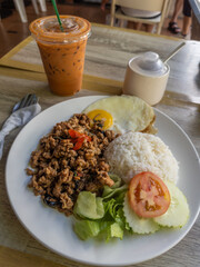 Authentic Thai street food meal featuring a plate of flavorful Pad Kra Pao with minced pork, a fried egg, and steamed rice, accompanied by a refreshing iced Thai milk tea, served on a wooden table in