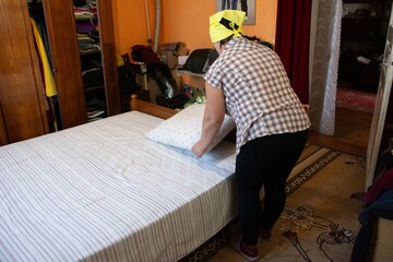 Woman makes a bed in a sunny bedroom with a large closet. She wears a yellow bandana. A carpet is visible on the floor.