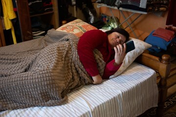 Woman in red pajamas uses her phone while lying in bed under a gray blanket, in a cozy bedroom with wooden bedframe and wardrobe.