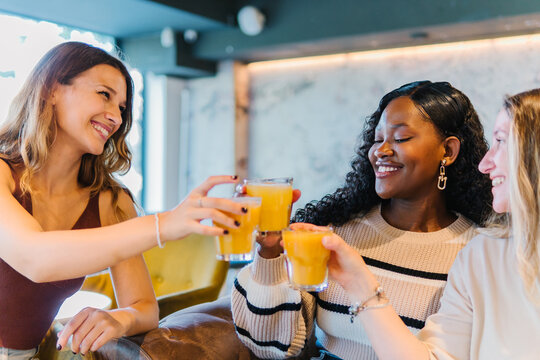 Diverse group of smiling women friends toasting with orange juice glasses, celebrating friendship and enjoying a cheerful brunch gathering in a modern cafe setting