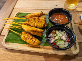 Delicious Thai chicken satay skewers, perfectly grilled and served on a banana leaf with authentic peanut sauce and refreshing Ajaat cucumber relish on a rustic wooden table.