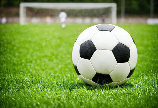 a single soccer ball on a lush green grass field with a blurred goal and player in the background, ready for a game or practice.
