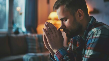 A young Caucasian man with a beard sits in a cozy living room, hands clasped in prayer. Soft lighting creates a warm atmosphere.