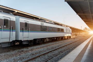 A train rests on the tracks at a quiet station as the sun rises, casting warm light and creating a serene morning scene. The station is empty, evoking a sense of tranquility