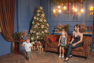 Elegant mother and her two young children posing on a leather sofa next to a glamorous gold-decorated Christmas tree, celebrating the holiday season in a stylish dark blue interior