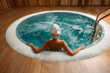 Back view of woman in towel turban relaxing in round jacuzzi with bubbling turquoise water, arms resting on edge, serene spa ambiance, wooden interior, tranquility, cleansing, healthy lifestyle