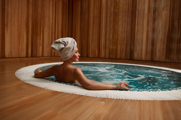 Back view of woman in towel turban relaxing in round jacuzzi with bubbling turquoise water, arms resting on edge, serene spa ambiance, wooden interior, tranquility, cleansing, healthy lifestyle