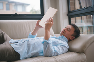 Portrait of asian man lying on a couch or sofa while using digital tablet in living room